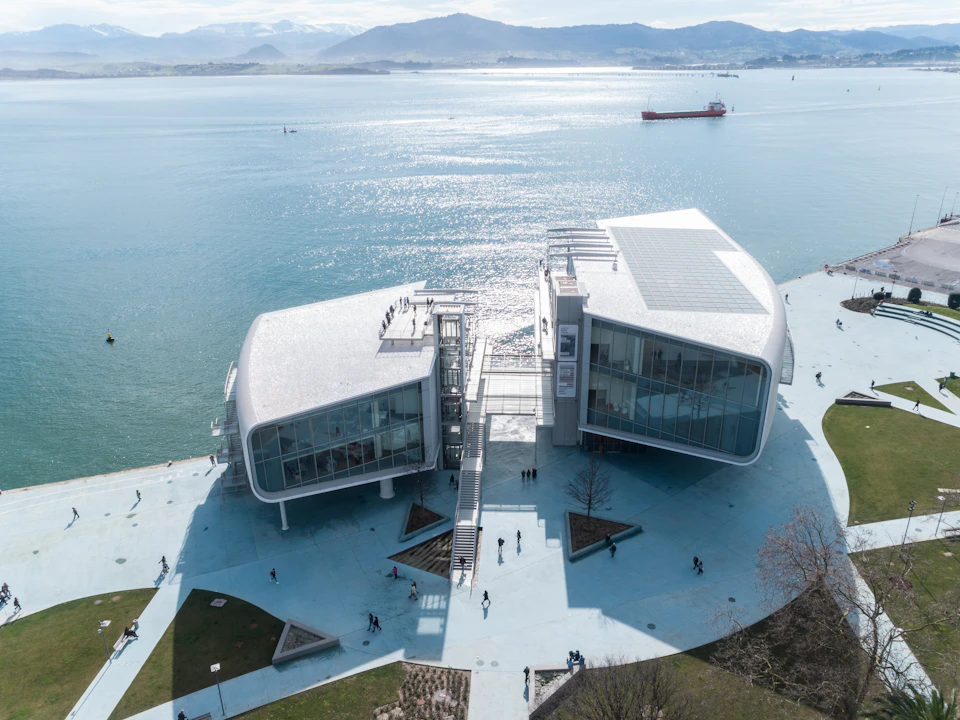 A modern, split building with large glass windows by a waterfront, people walking on the paved area, mountains in background.