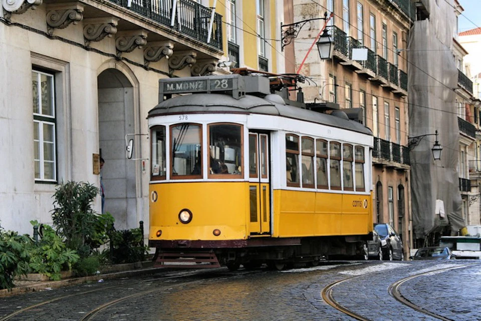 Yellow tram navigating through a narrow, cobblestone street lined with historic buildings and balconies in an urban setting.