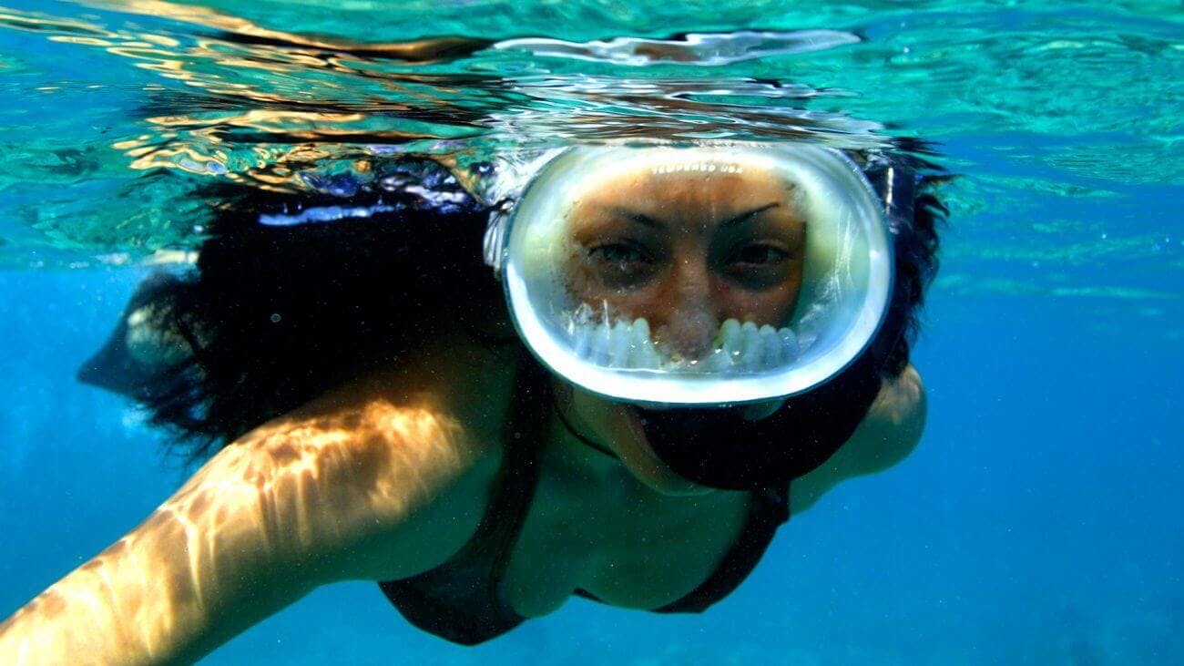 Persona que fa esnórquel sota l'aigua amb mascareta integral i aigua blava clara al seu voltant.