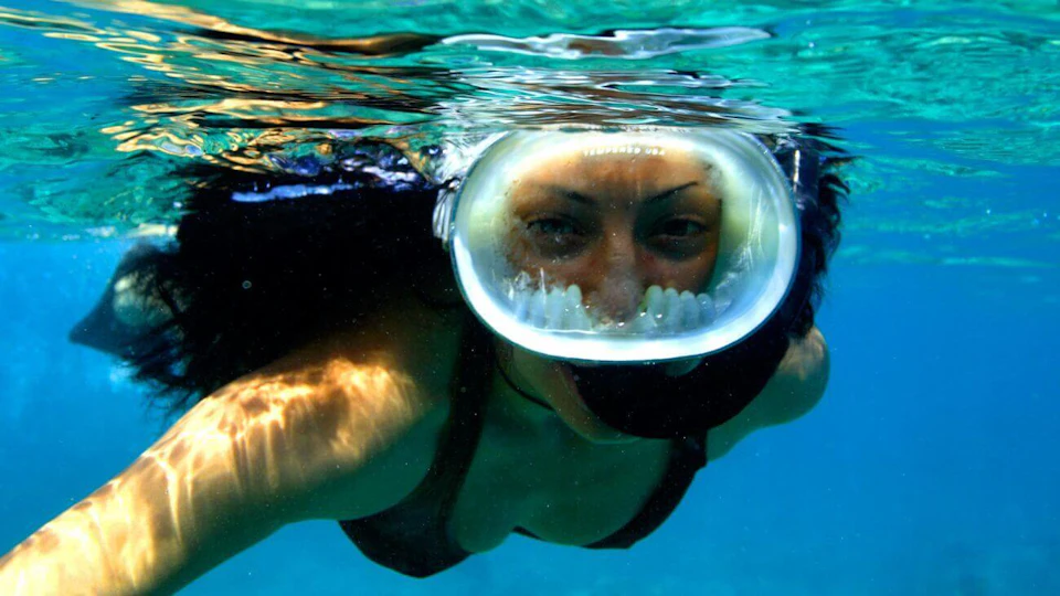 Person snorkeling underwater wearing a full-face mask, with clear blue water surrounding them.