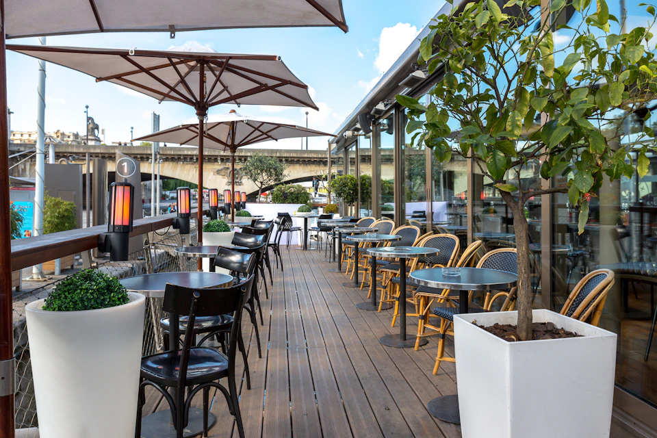 Outdoor cafe with wooden decking, tables, chairs, umbrellas, potted plants, and a view of a bridge in the background.