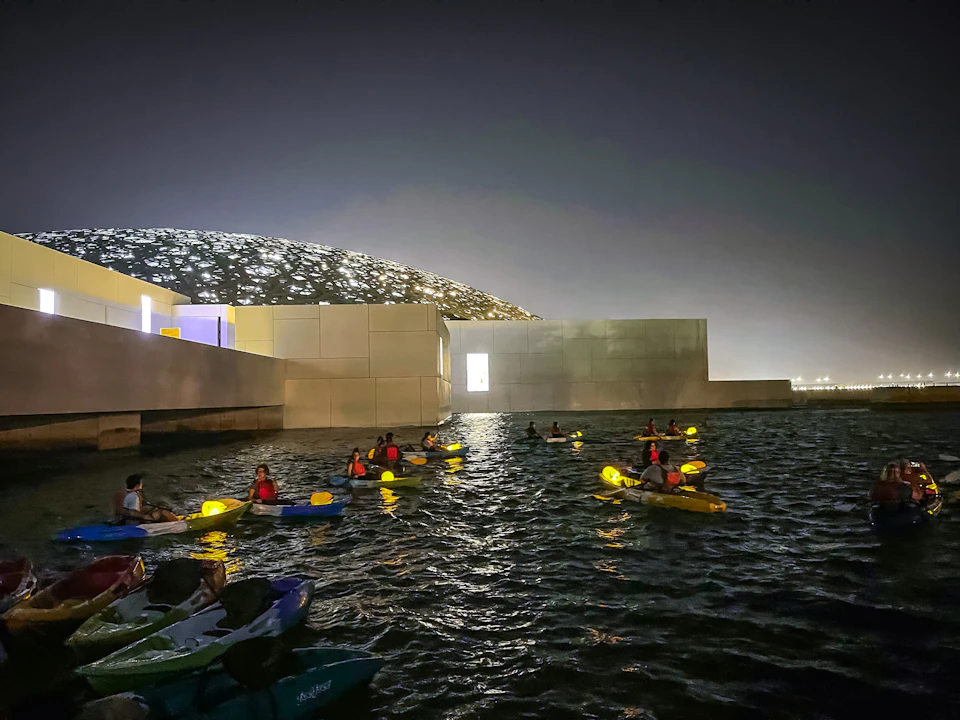 People kayaking in a dark, illuminated water area near a modern building with a latticed, dome-shaped roof in the background.