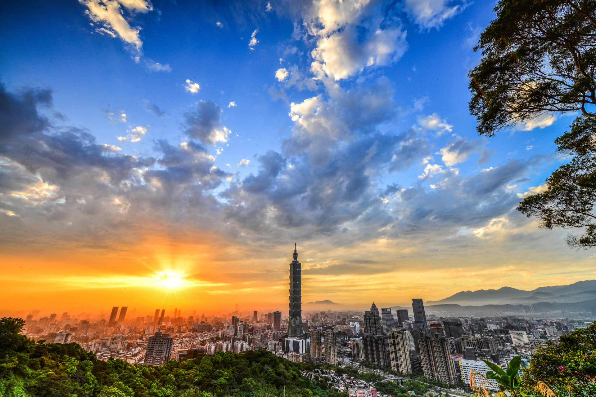 Cityscape at sunrise with a tall tower in the center, surrounded by skyscrapers, hills, and a vibrant sky with clouds.