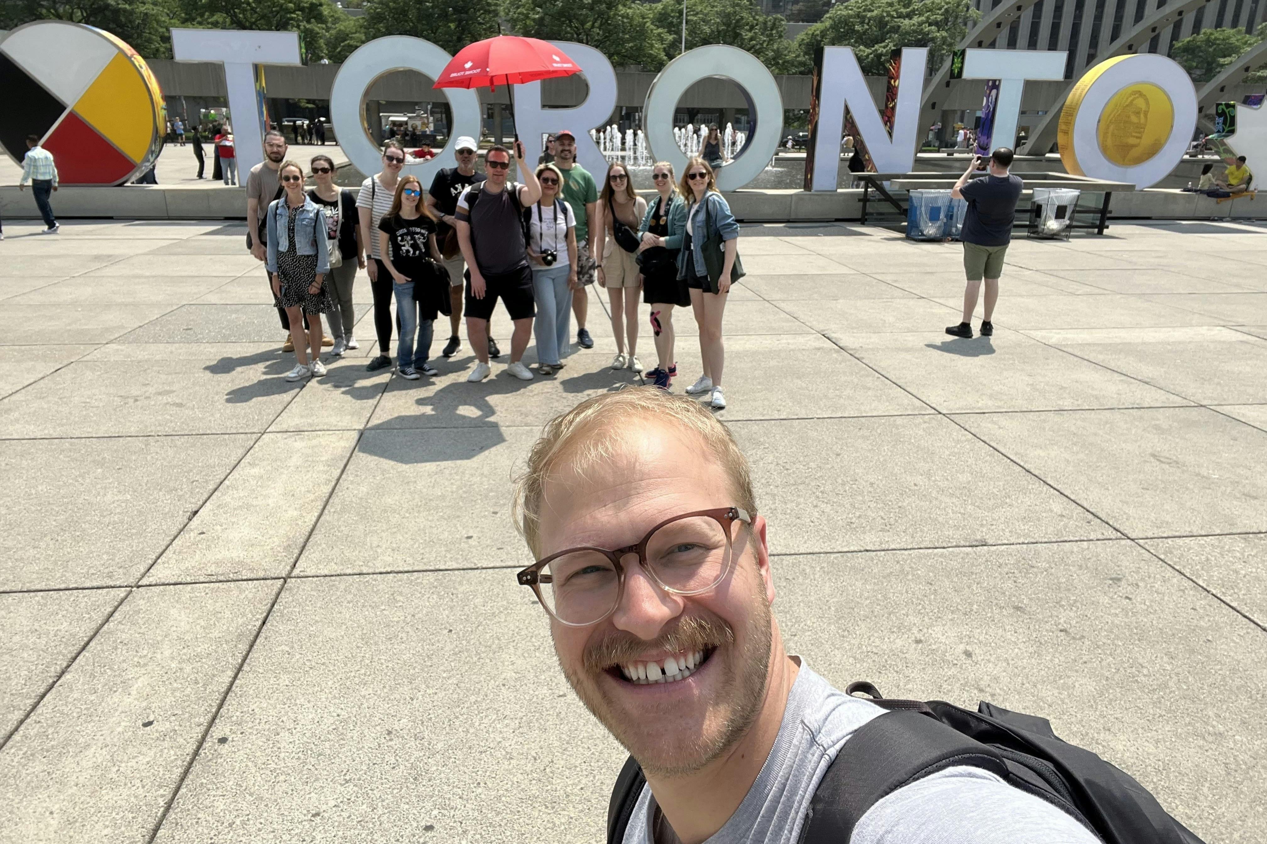 一群人在广场上的大型“多伦多”信件前摆姿势，前景中有一个人自拍。
