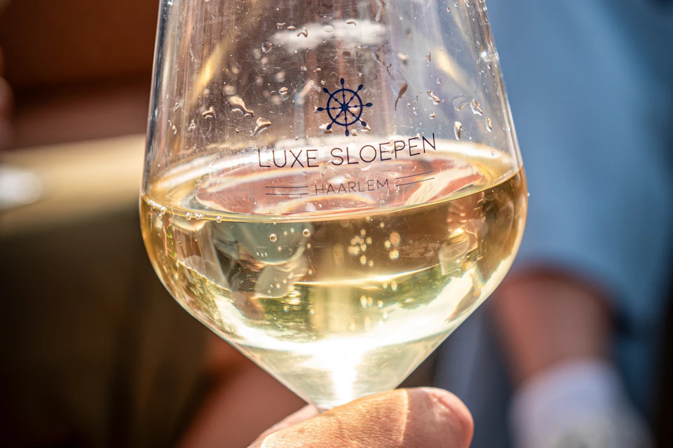 A close-up of a hand holding a wine glass with white wine, showing the text 'LUXE SLOEPEN HAARLEM' printed on the glass.