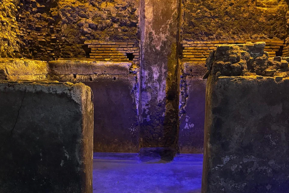 An ancient stone structure illuminated with yellow and blue lights, featuring a central wall and a water-filled foreground.