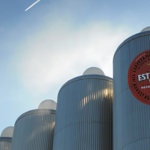 Höga industrisilor i silver med en stor Estrella Damm-logotyp mot en klar himmel och en kontrail från ett flygplan.