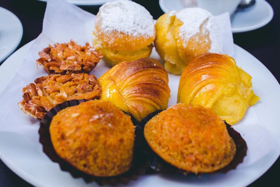 Assorted pastries on a plate, including powdered sugar-topped buns, croissants, and flaky, crumbly desserts.
