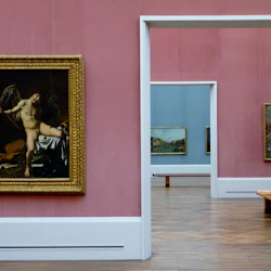 Konstgalleri med rosa och blå väggar som visar inramade målningar. En dörröppning avslöjar mer konstverk och en träbänk i nästa rum.