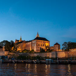 Ett upplyst slott med stenmurar och torn sitter på en kulle bredvid en strandpromenad under en klar kvällshimmel.