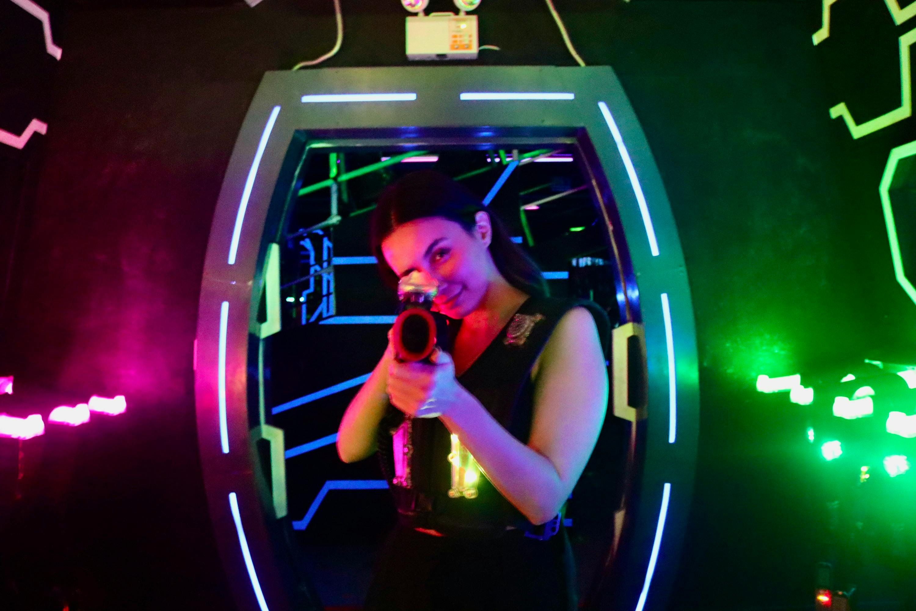A person standing in a neon-lit environment, holding a laser tag gun and wearing a vest, smiling towards the camera.
