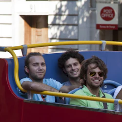 Fyra män på en dubbeldäckare ler mot kameran. Man håller en kamera, medan en "Post Office" -skylt syns i bakgrunden.