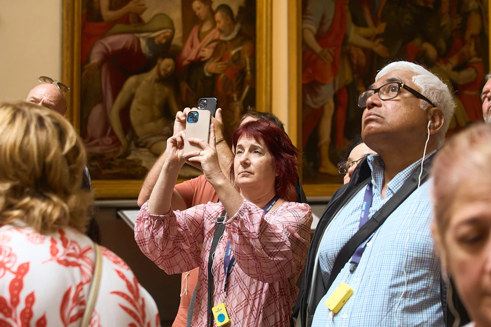 A group of people in a museum, some observing paintings on the wall, and a woman taking a photo with a smartphone.