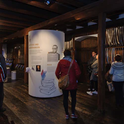 Människor observerar uppvisningar av historiska svärd och informationspaneler i en svagt upplyst utställningshall i trä.