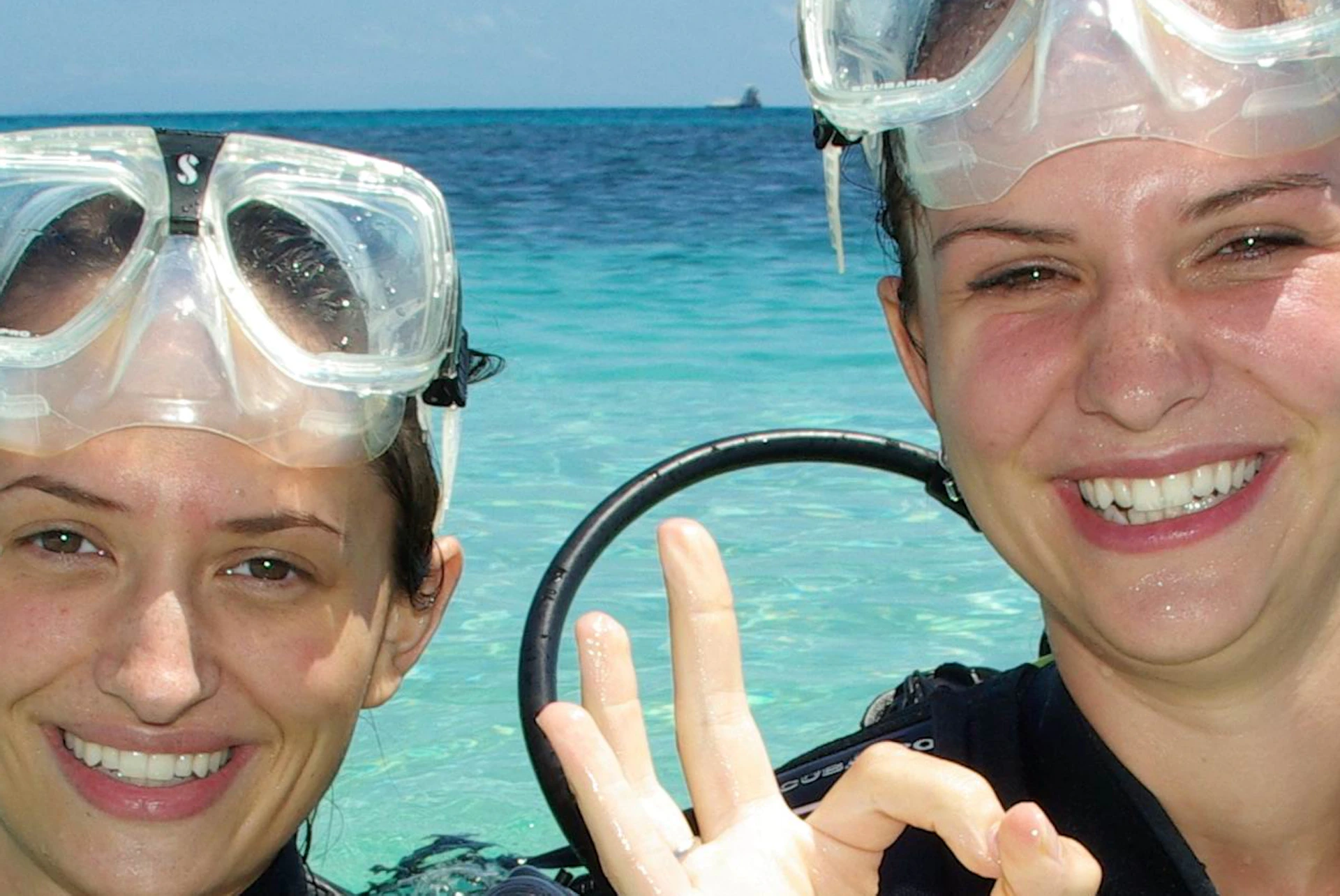 Two scuba divers in shallow, clear ocean water, smiling and making the 'OK' sign, with boats and blue sky in the background.