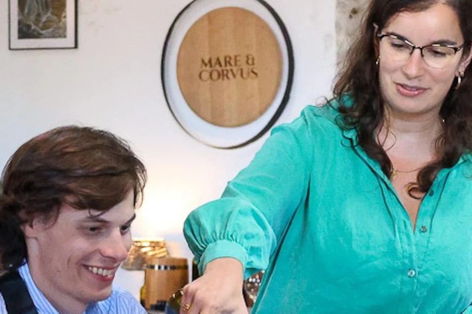A group of people seated at a table while a woman pours wine into glasses. The setting is indoors with a cozy atmosphere.