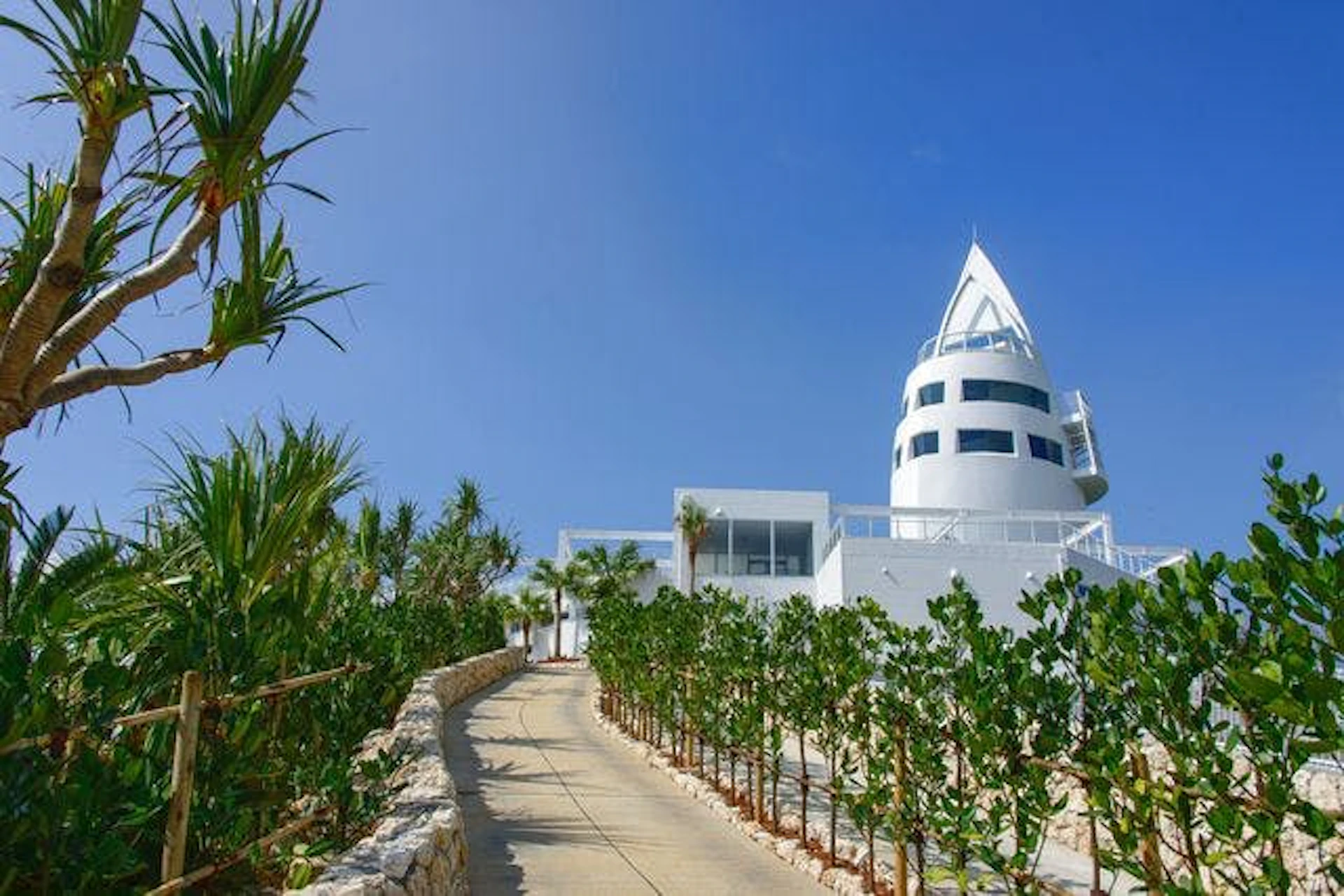 A white tower with a unique pointed roof stands amid lush greenery along a winding pathway under a clear blue sky.