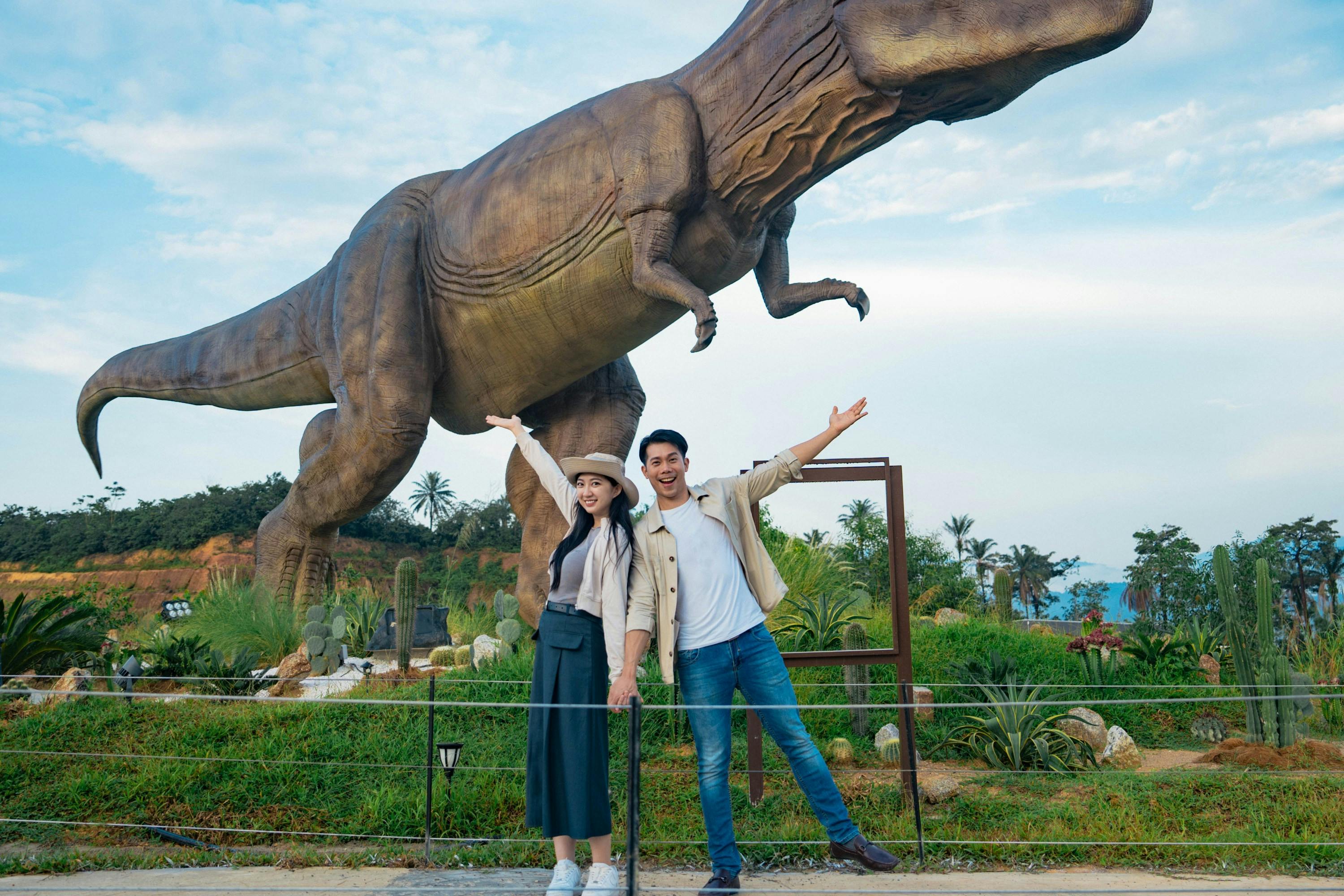 Two people posing with arms raised in front of a large dinosaur statue in an outdoor park setting.