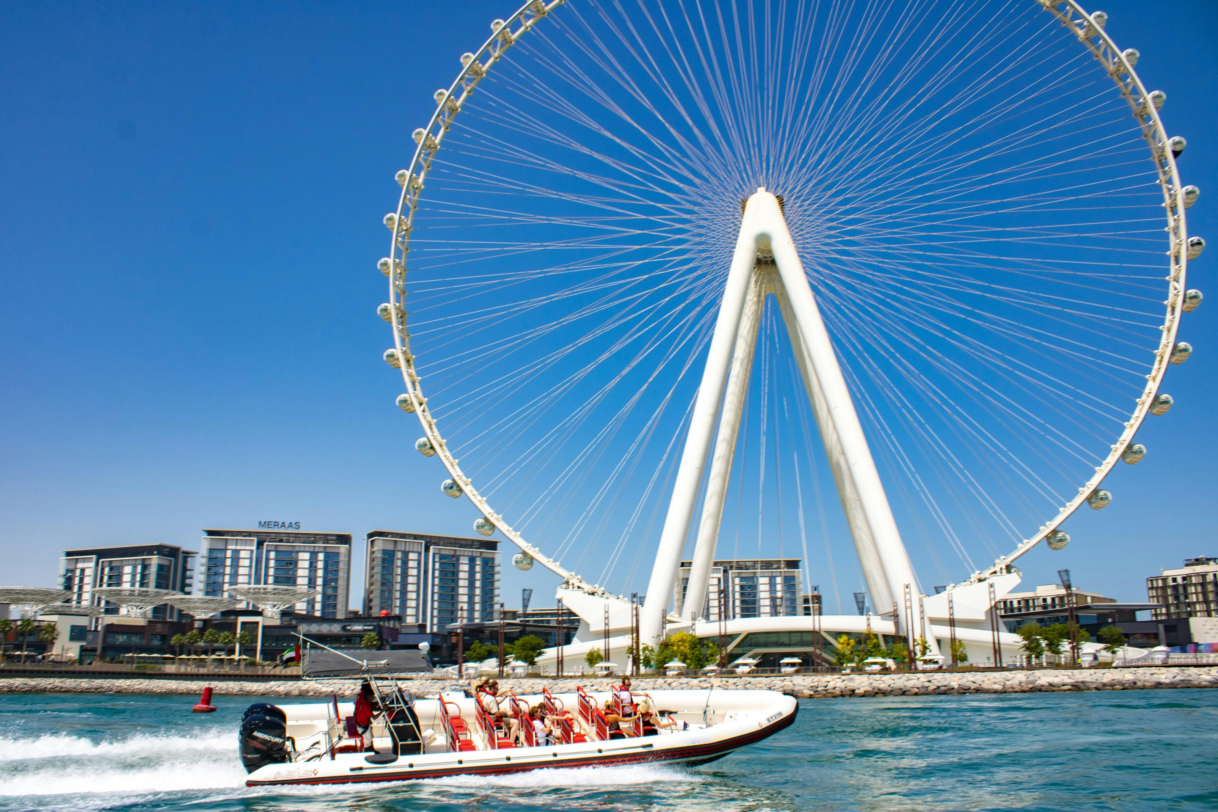 Před velkým ruským kolem a moderními budovami pod jasně modrým nebem je motorový člun na vodě s cestujícími.