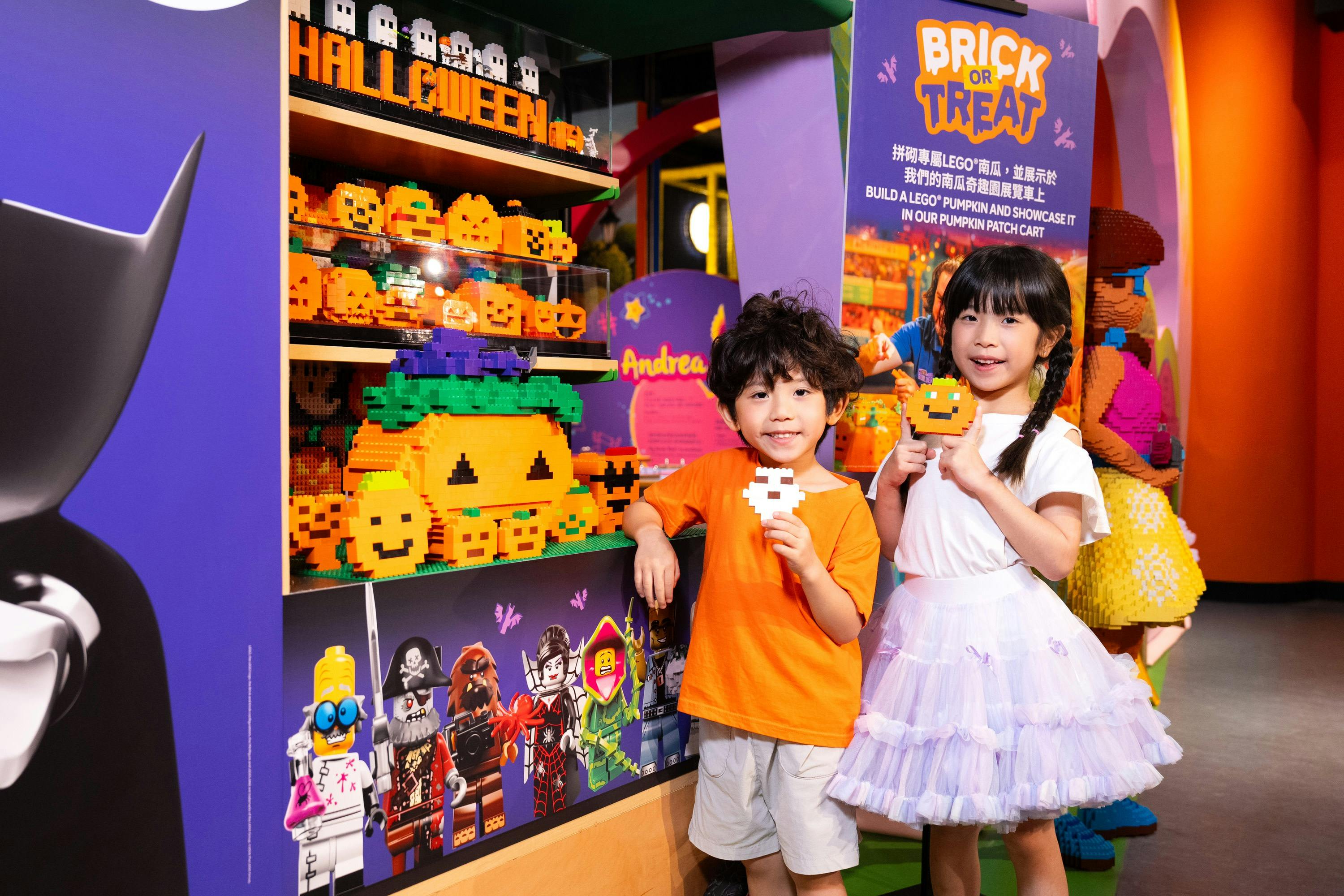 Two children smiling and holding LEGO Halloween-themed creations in a colorful store with decorations and LEGO displays.