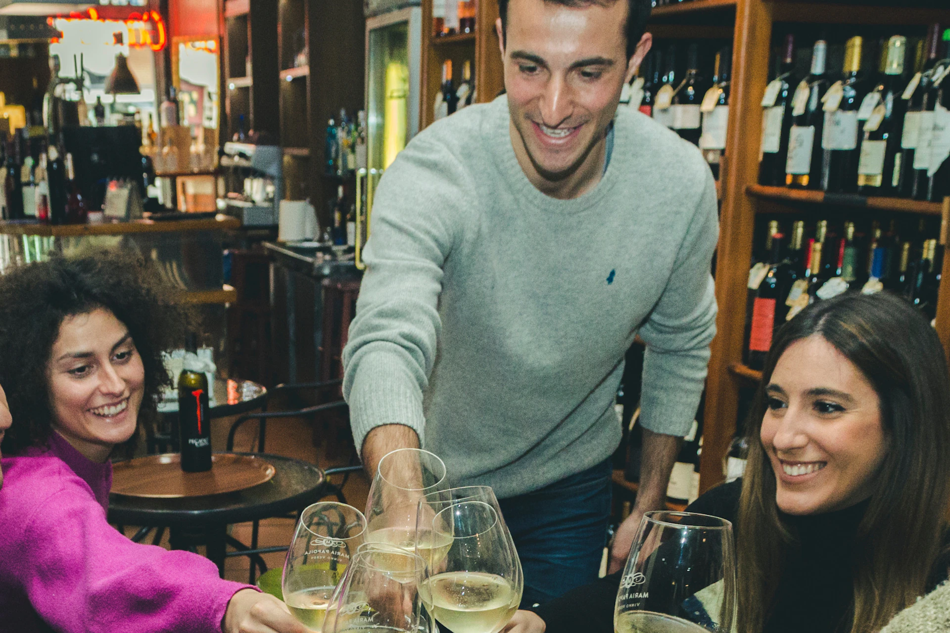 Five people are clinking wine glasses around a table with food, in a cozy, dimly-lit wine bar with shelves of wine bottles.