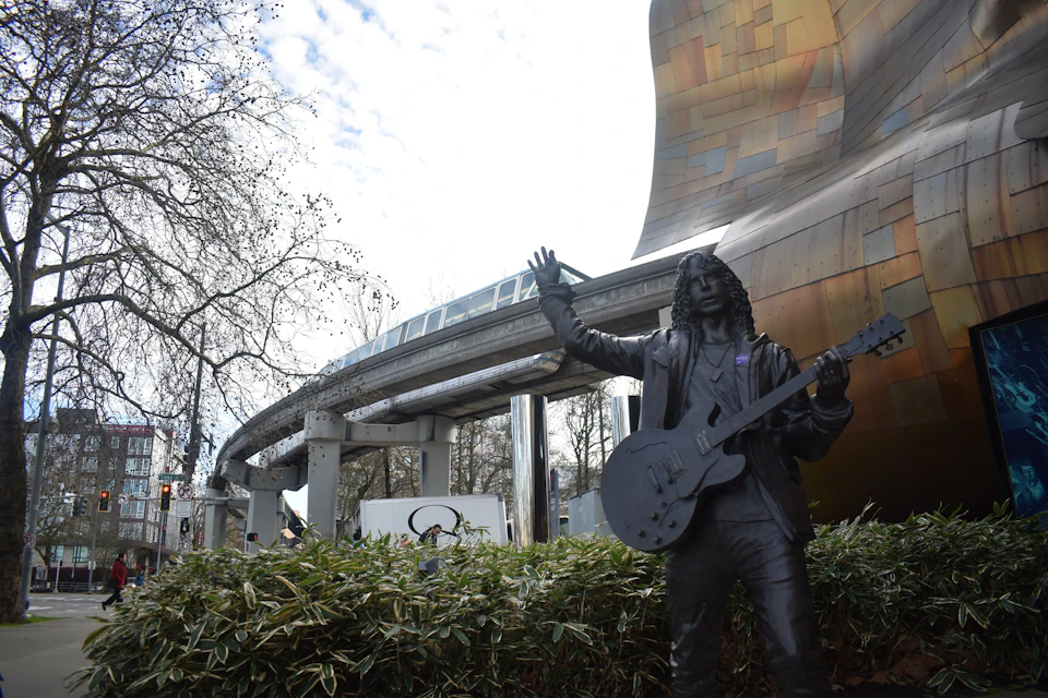 A statue of a man holding a guitar with an elevated monorail track in the background by a modern building and trees.
