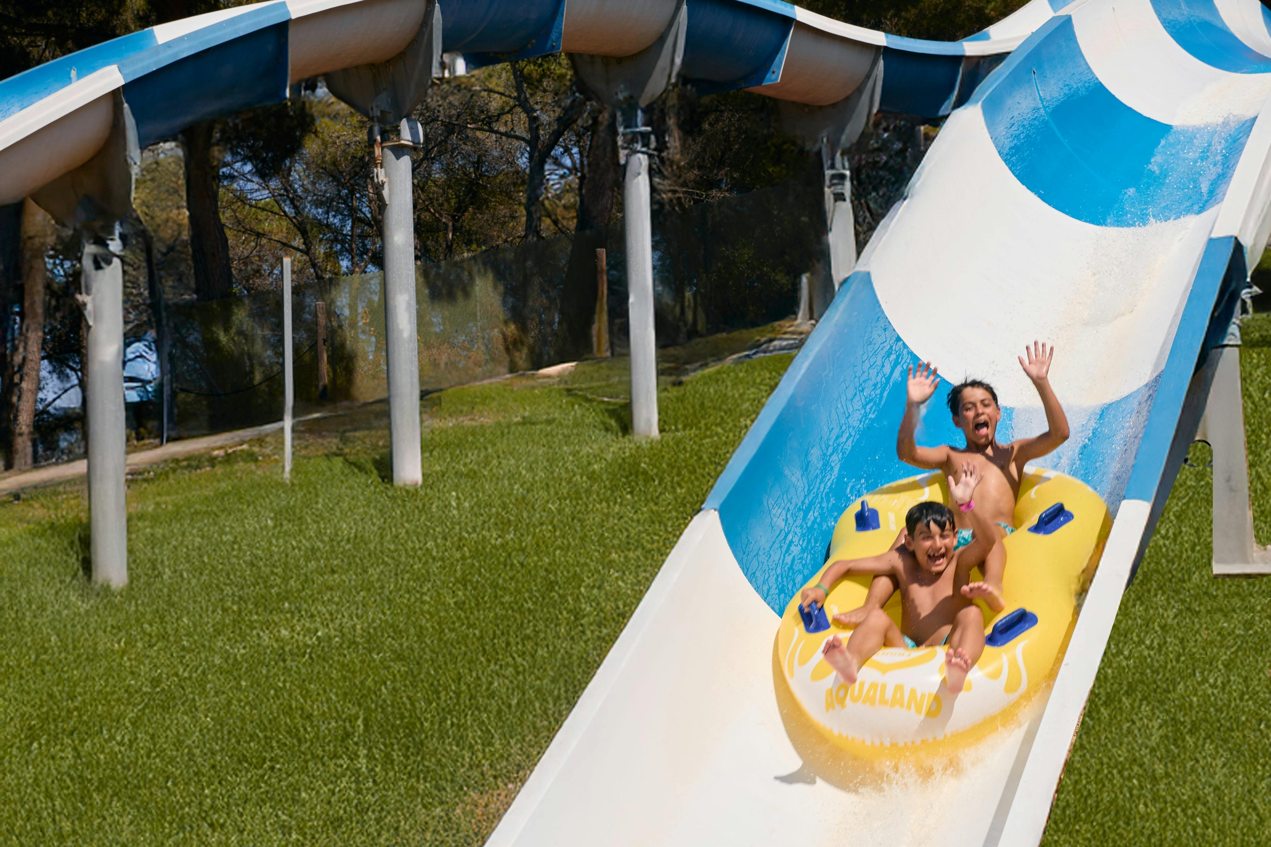 Two people riding a yellow inflatable down a blue and white water slide, smiling and raising their hands in excitement.