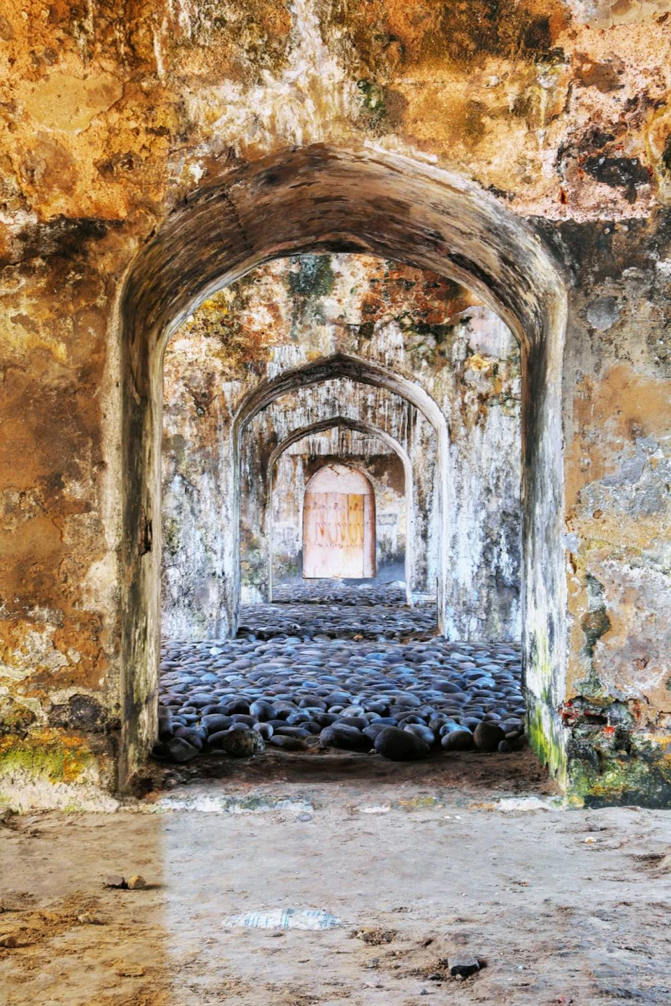 A series of weathered stone archways with a pebbled floor, leading to a closed wooden door in an old, textured building.
