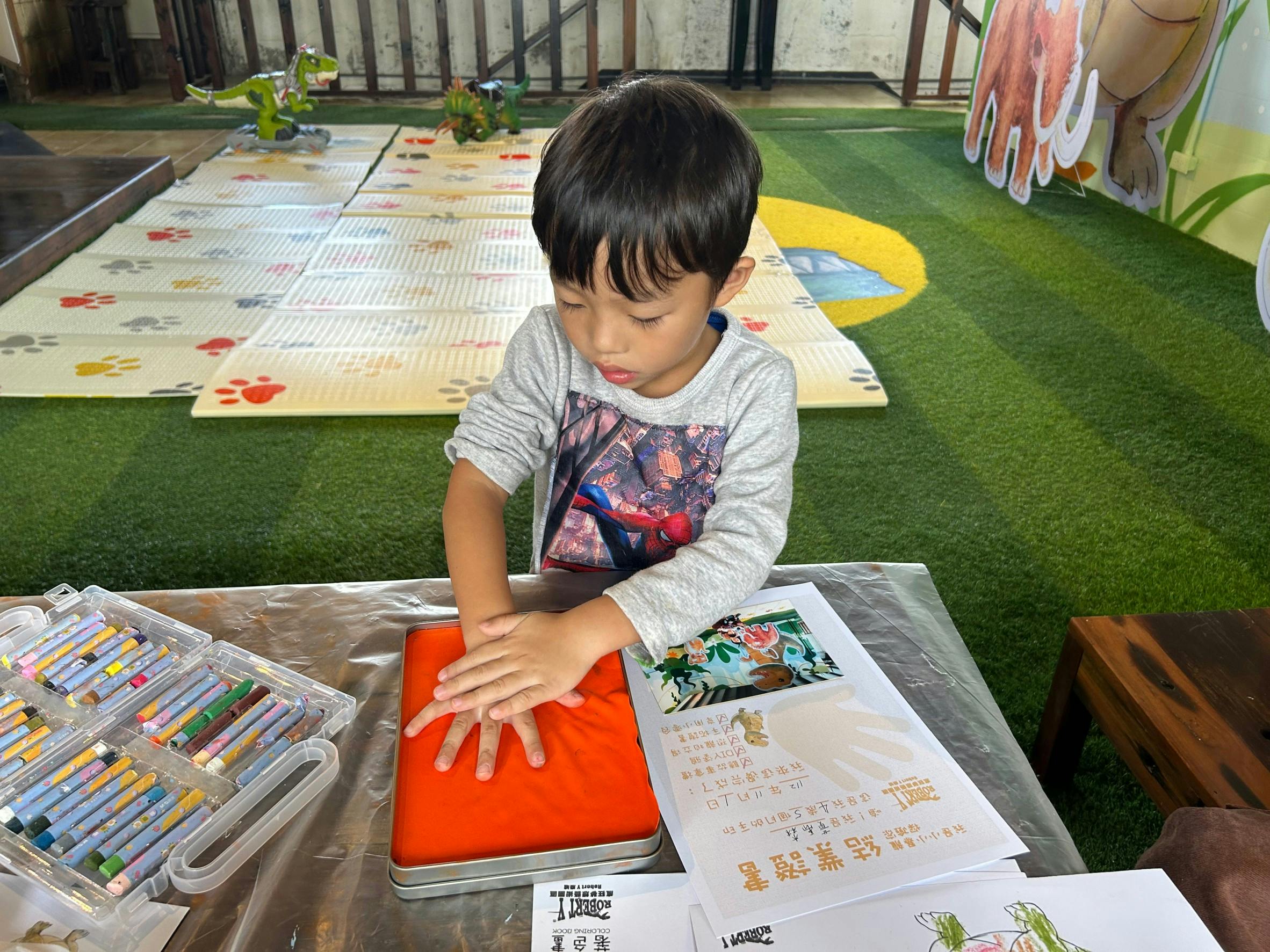 A young boy pressing his hands into red clay at a table with art supplies and paper in an indoor play environment.