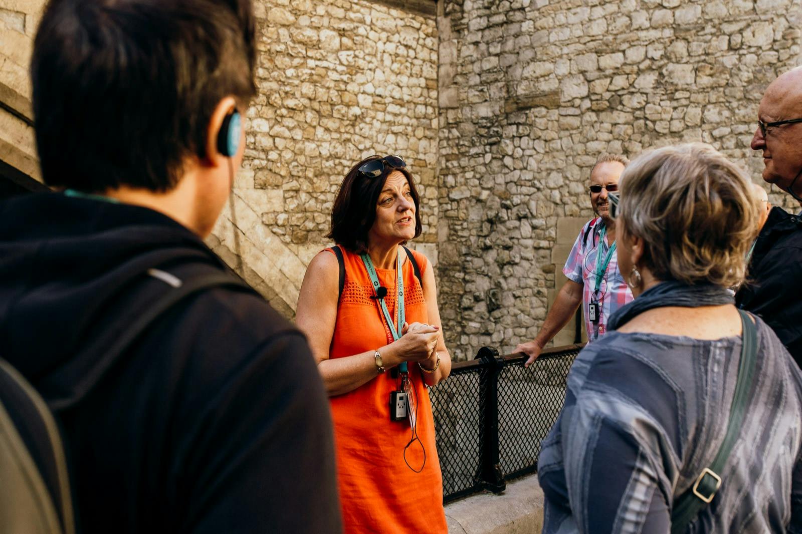 A tour guide in an orange dress talks to a group of people in front of a stone wall.