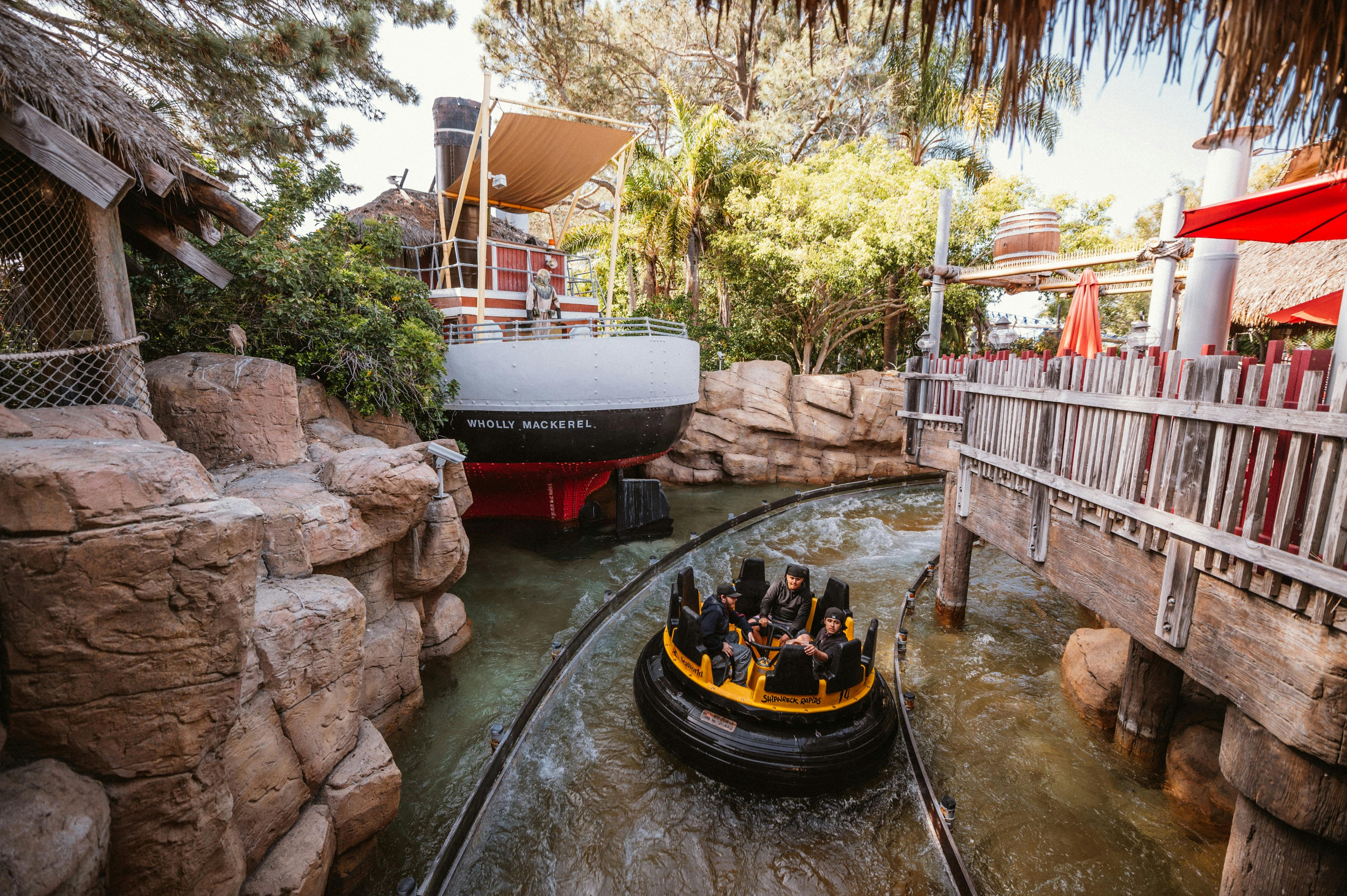 Οι άνθρωποι σε μια κυκλική βόλτα με σχεδία περνούν από μια βάρκα που ονομάζεται "Wholly Mackerel" σε μια βόλτα στο θεματικό πάρκο που περιβάλλεται από βράχους και δέντρα.