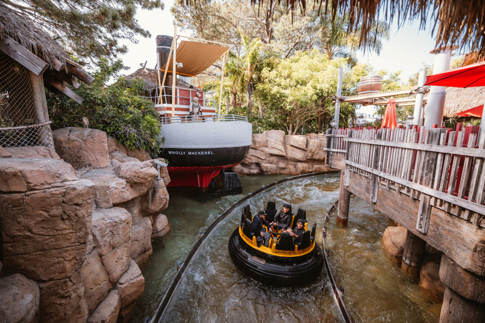 People in a circular raft ride pass by a boat named "Wholly Mackerel" in a theme park water ride surrounded by rocks and trees.