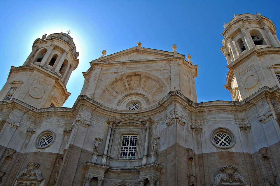 Sunlit facade of a grand, ornate stone cathedral with two towers against a clear blue sky.