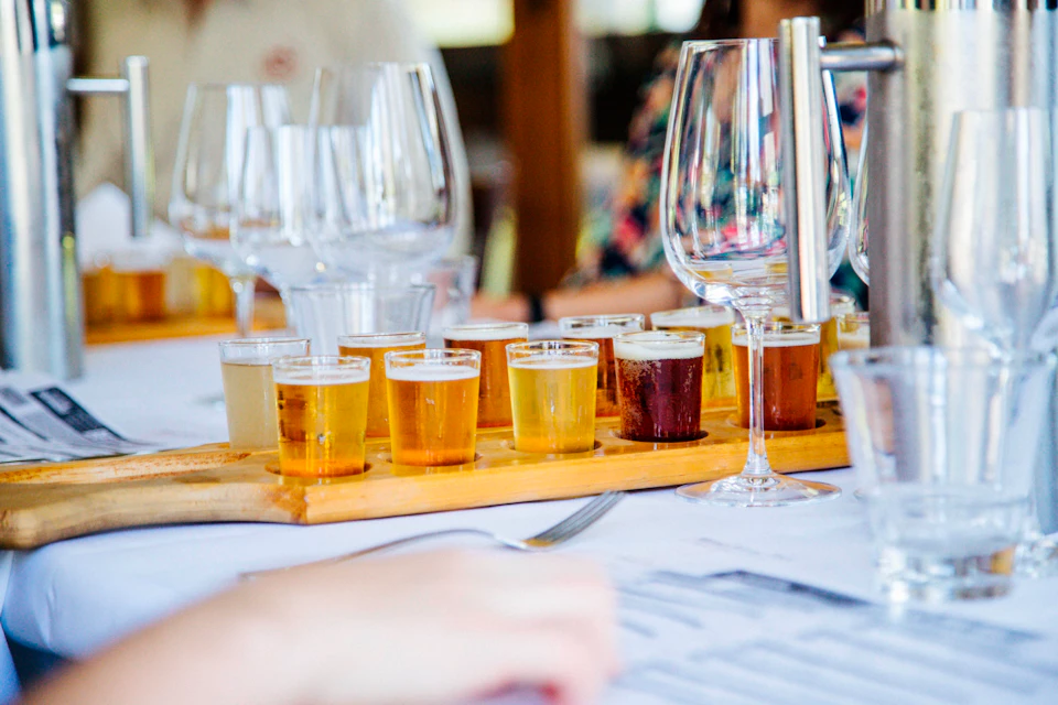 A flight of various beers in small glasses on a wooden tray, surrounded by wine glasses on a restaurant table.