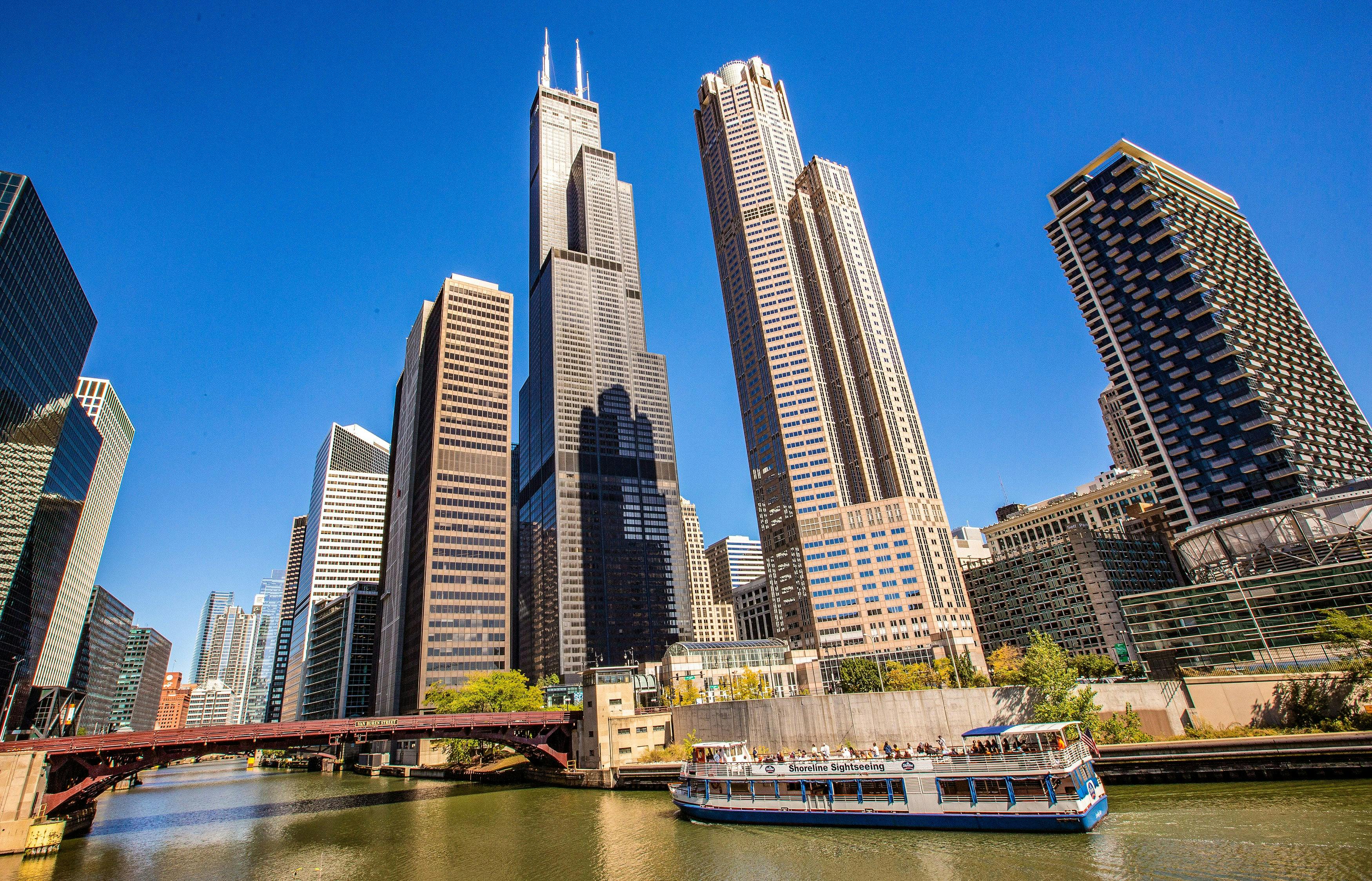 Skyscrapers, including a tall black one with twin antennas, along a river with a tour boat under a clear blue sky.