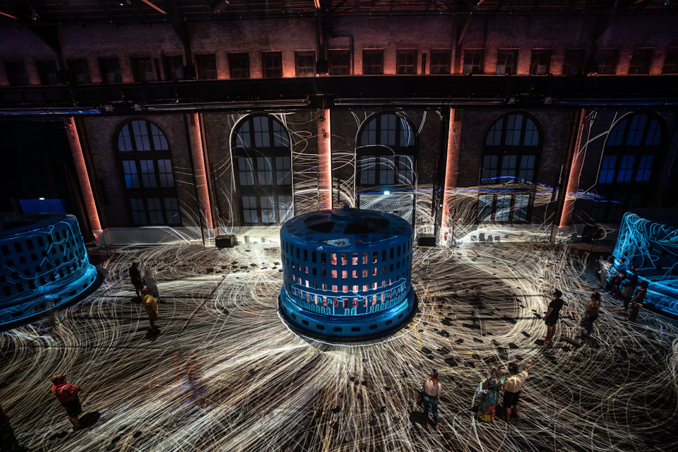 A large blue cylindrical installation emits light trails in a dimly lit industrial space with people observing.