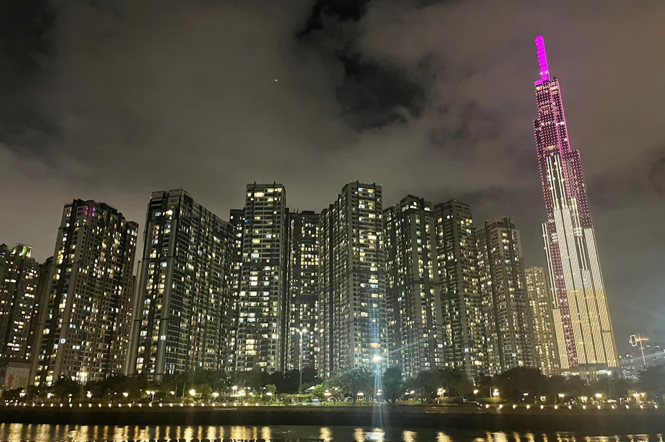 Tall city buildings lit up at night, with a river and trees in the foreground. A building with a pink-lit spire is on the right.