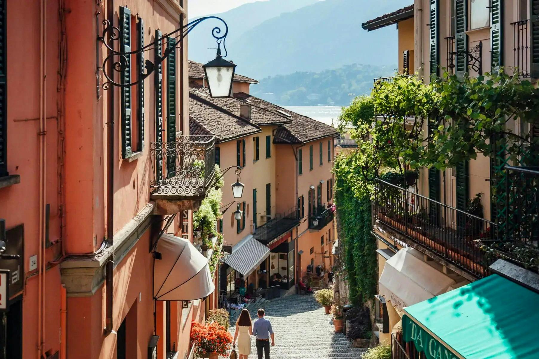 A picturesque cobblestone street lined with charming buildings, greenery, and a couple walking downhill towards a lake.