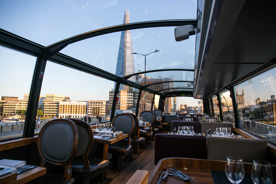 Interior of a glass-roofed restaurant bus, set dining tables, view of The Shard skyscraper and London cityscape outside.