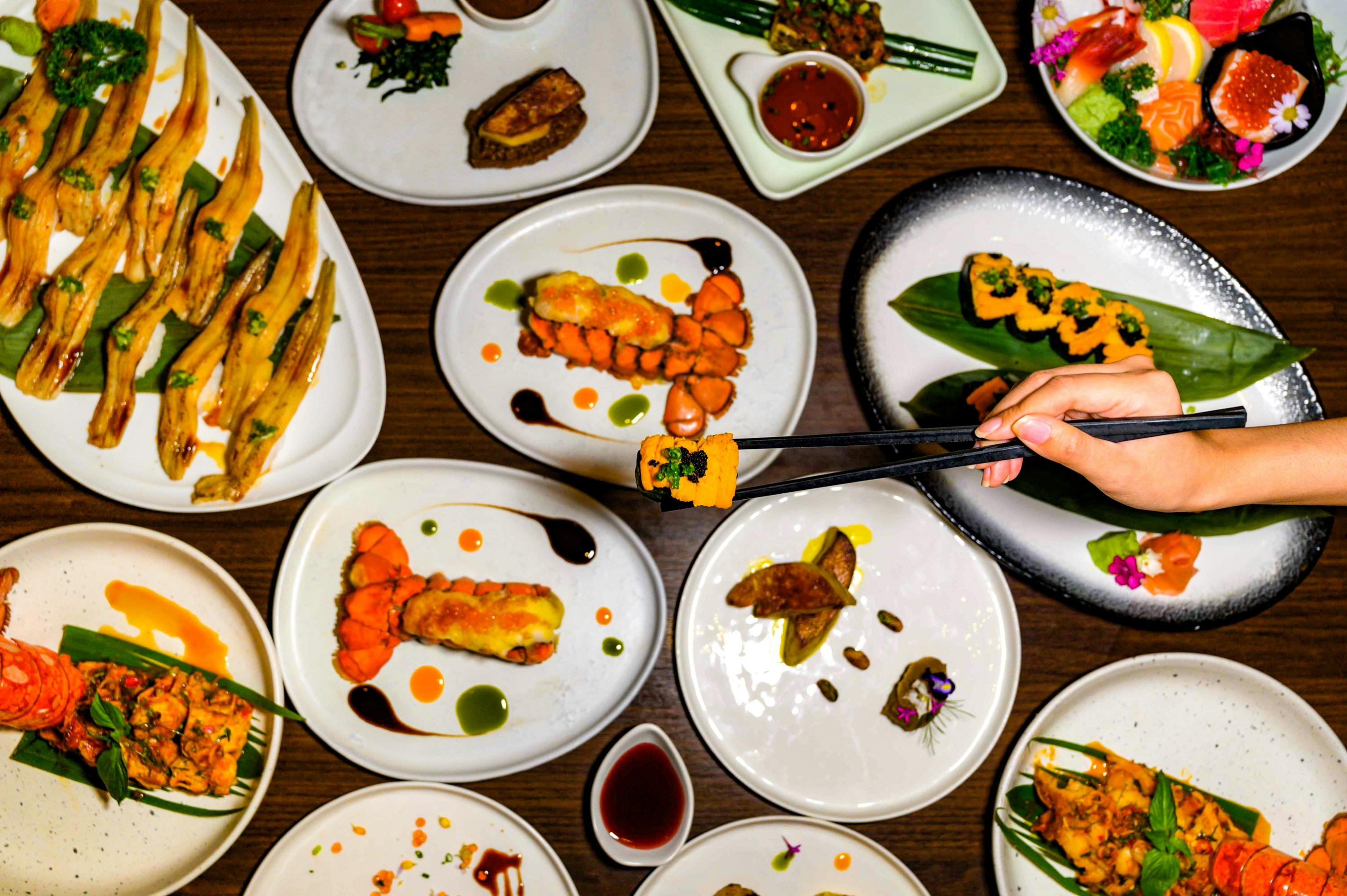 A variety of plated dishes on a wooden table, with a hand using chopsticks to pick up food from one dish.