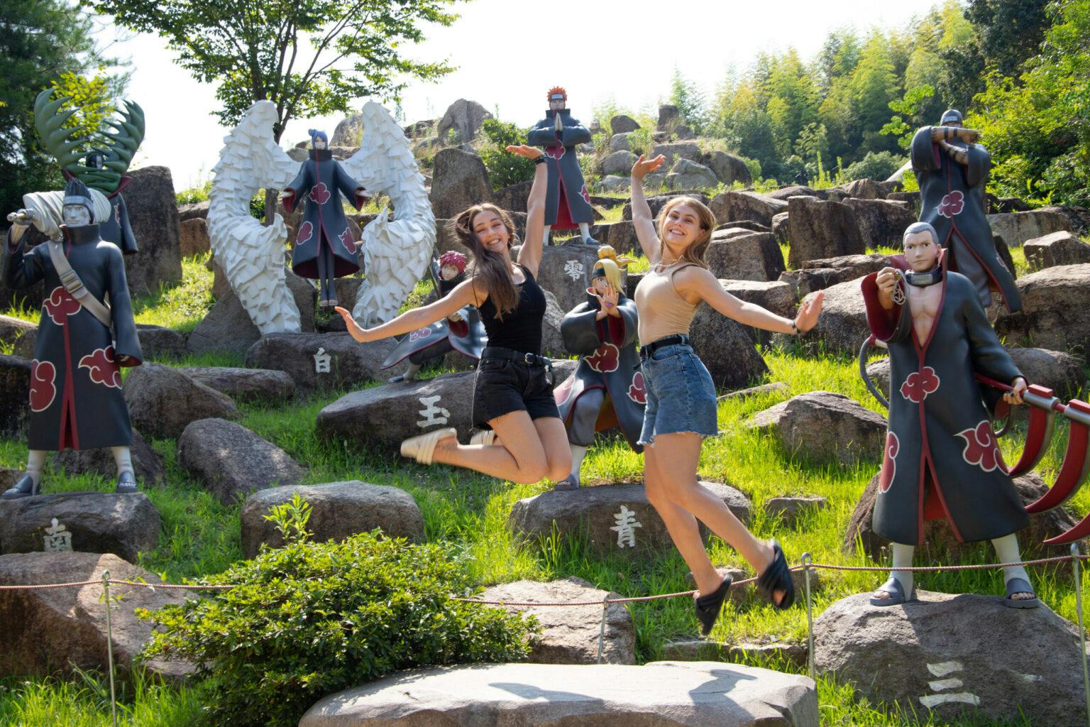 Two people jumping and posing energetically near life-sized anime character statues in an outdoor park setting.