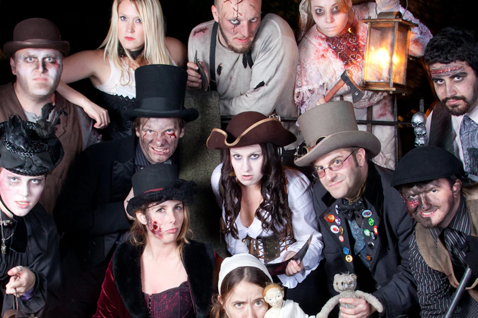 A group of people in vintage costumes holding lanterns and tools, posing on grass, with dark trees and an orange sky in the background.
