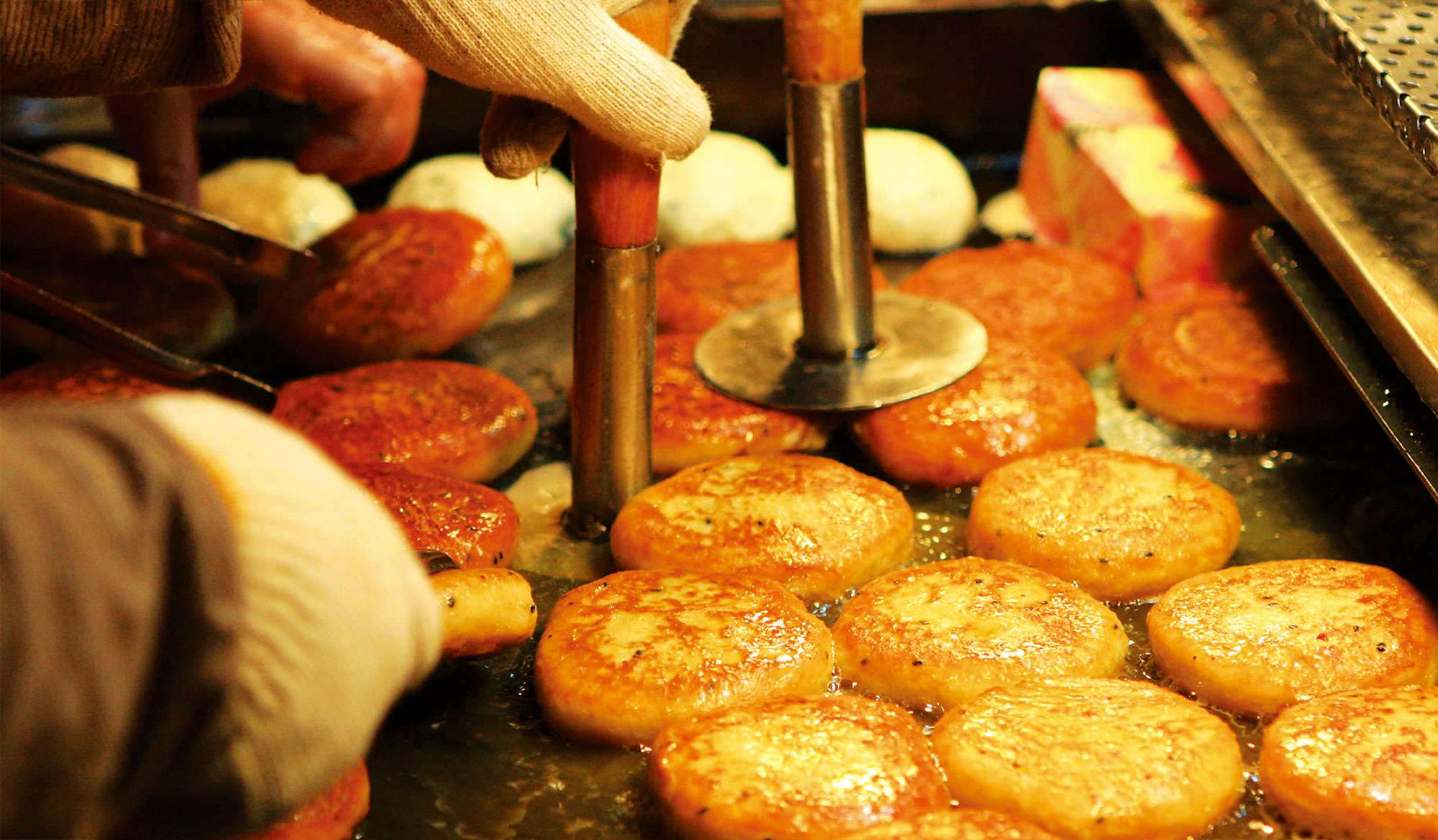 Dłonie z narzędziami spłaszczającymi porcje ciasta na patelni podczas smażenia, tworząc złocistobrązowe okrągłe przekąski.