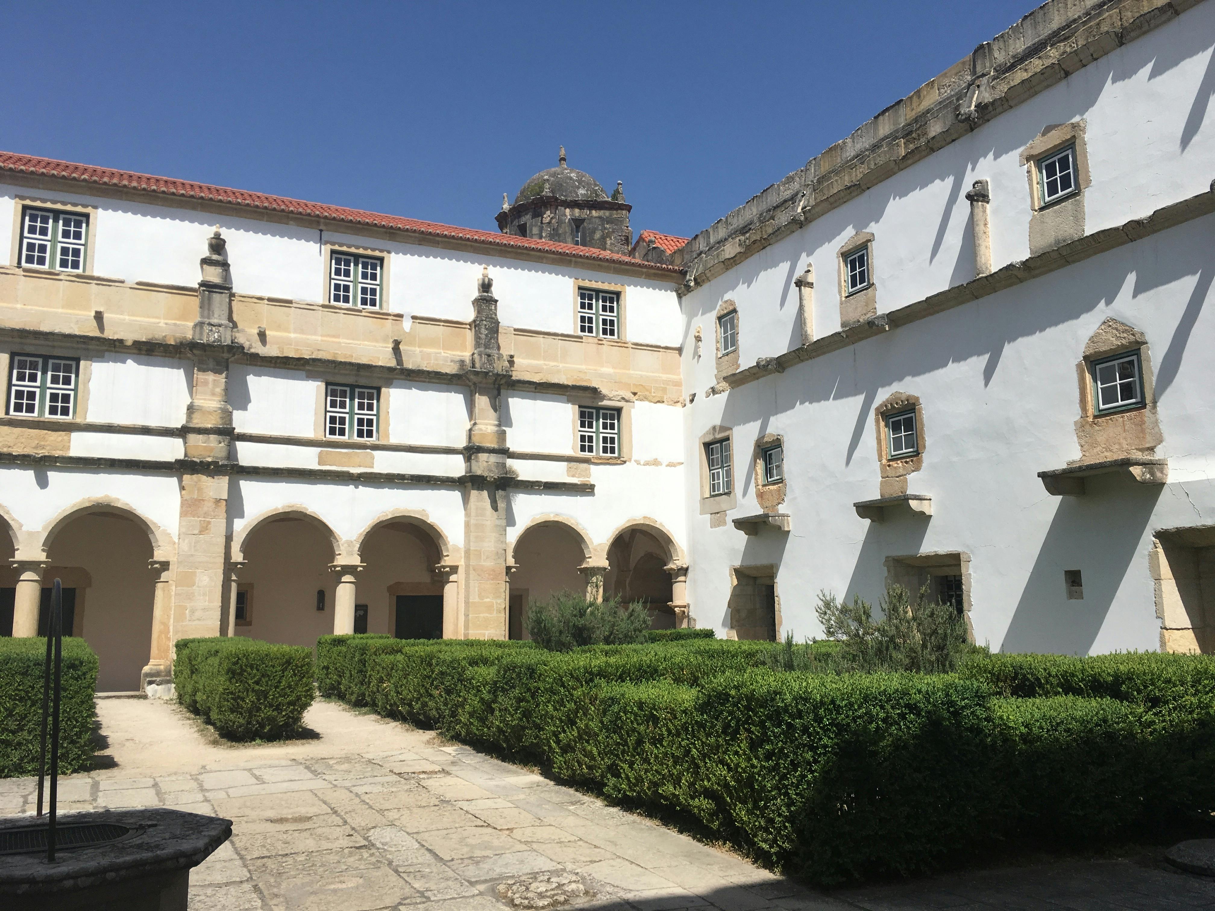 澄んだ青空の下、アーチ型の戸口と窓がある2階建ての歴史的な建物に囲まれた、きれいに整えられた生垣のある中庭。