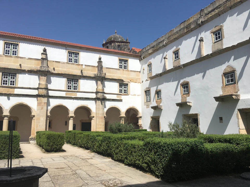 A courtyard with neatly trimmed hedges, surrounded by a two-story historical building with arched doorways and windows, under a clear blue sky.