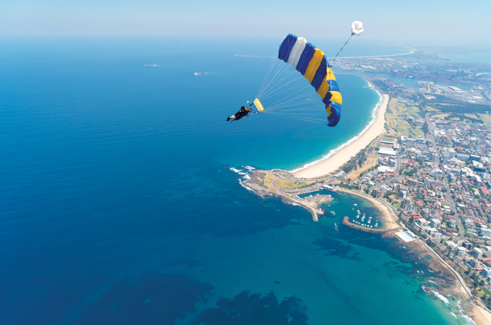 Skydiver descending with a blue, yellow, and white parachute over a coastal landscape with blue ocean, beach, and buildings.