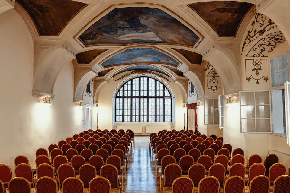 A hall with rows of red chairs, an arched window at the front, and a ceiling adorned with artwork.