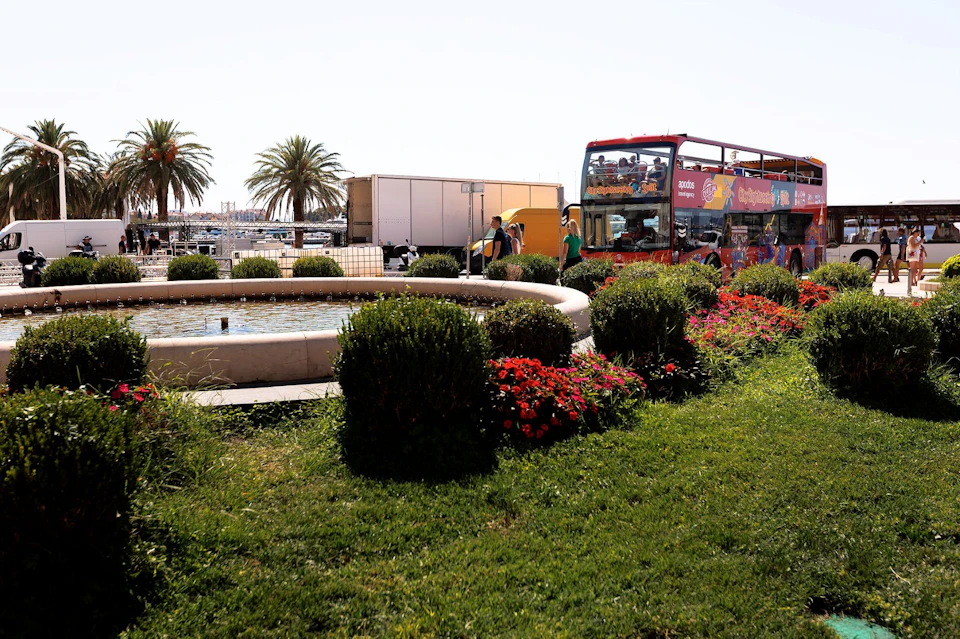 A red double-decker tour bus parked near a circular fountain with flowers and palm trees in the background.