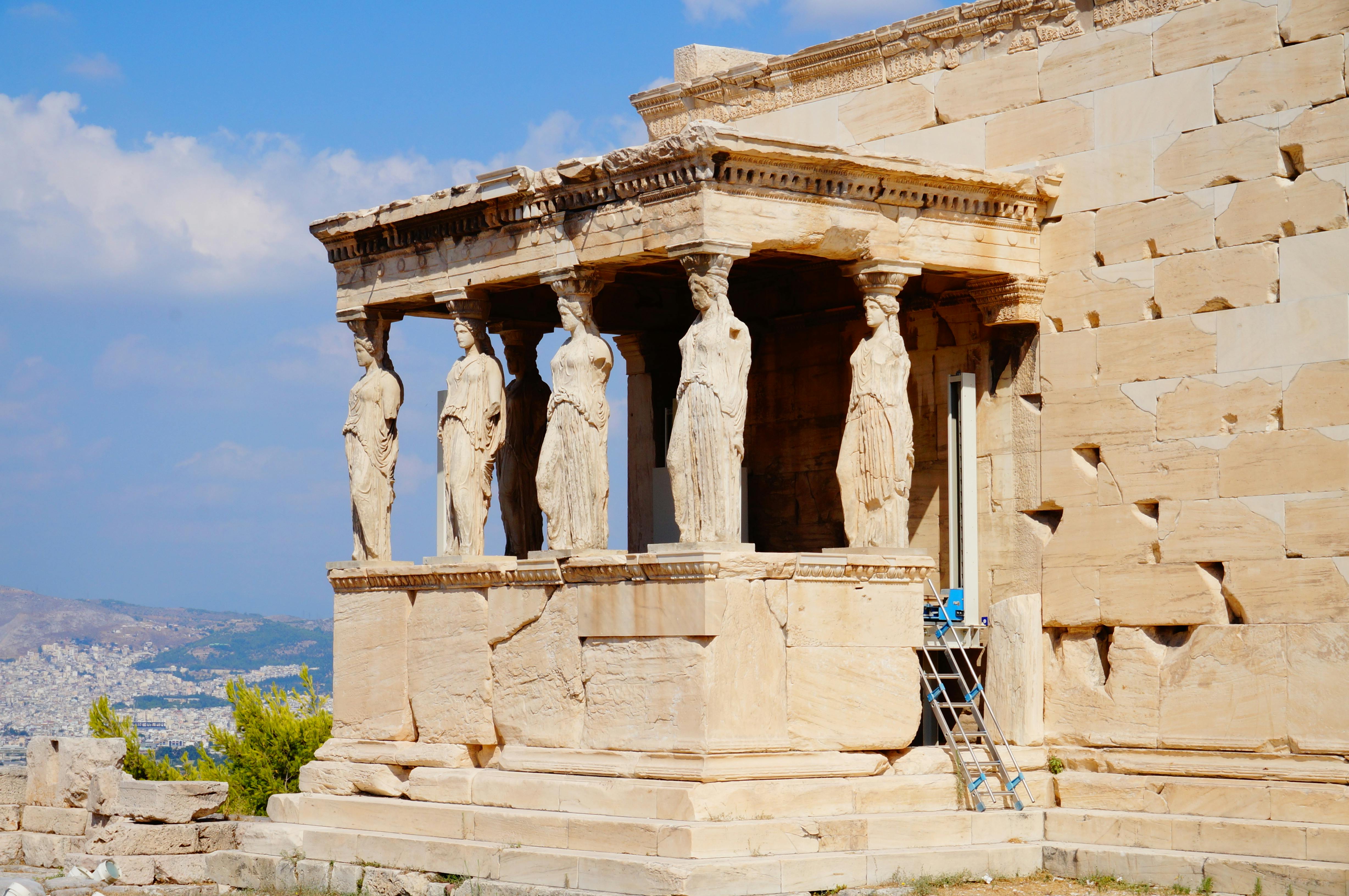 Ancient Greek temple structure with six caryatid columns, each carved as a draped female figure. Blue sky and ladder visible.