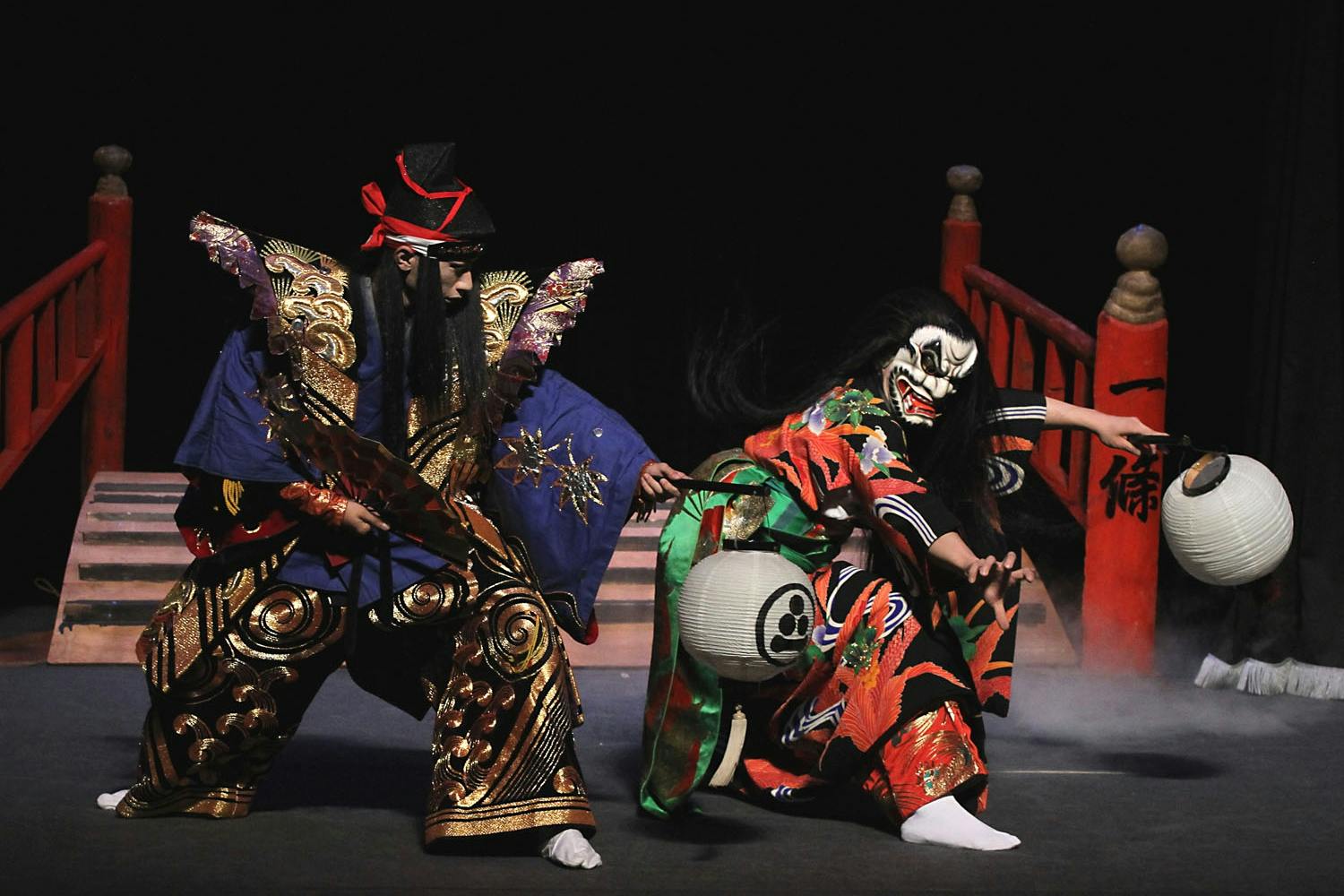 Two performers in elaborate traditional costumes and masks engaging in a dramatic pose on a stage during a performance.